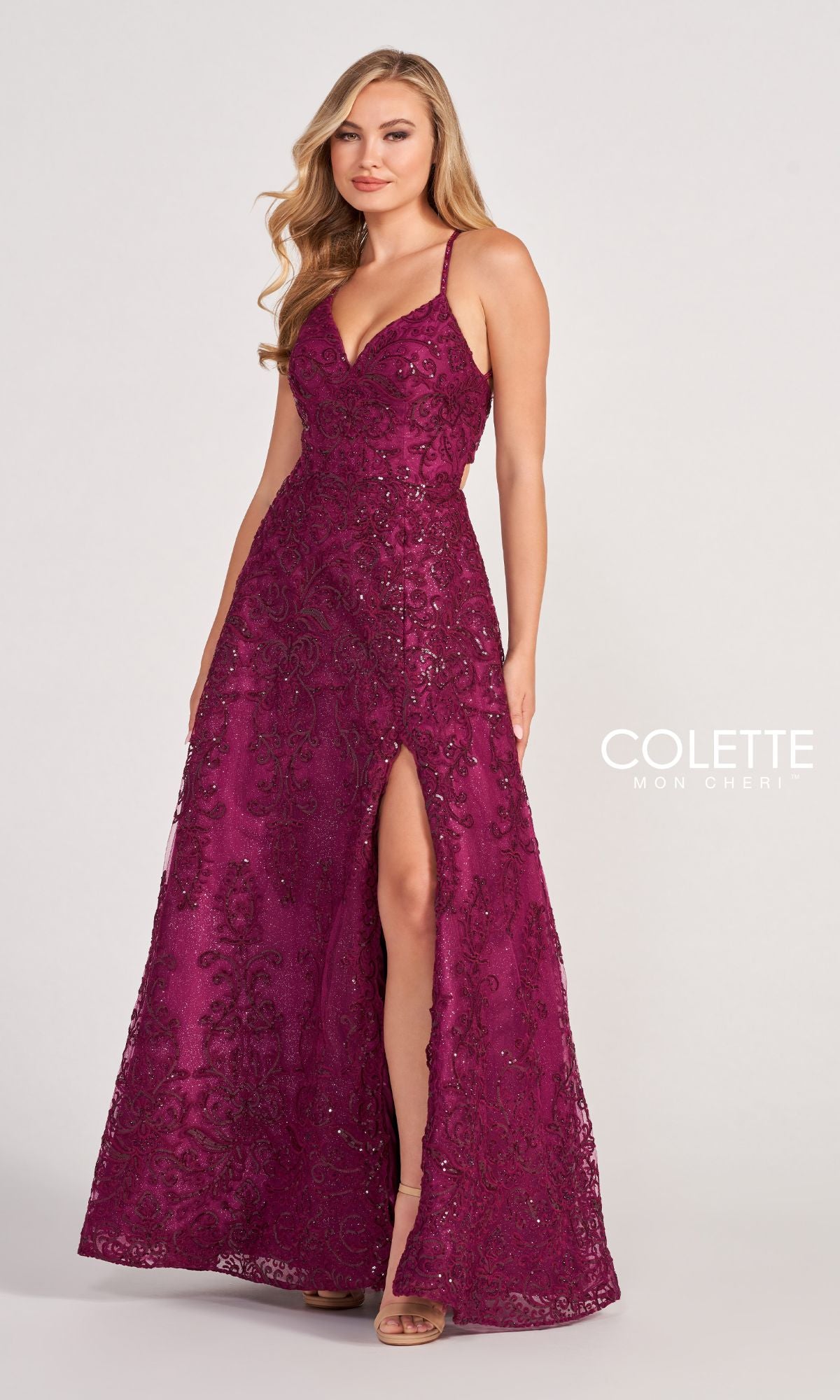 A woman stands against a plain light background wearing the Colette CL2028 Strappy-Back Lace A-Line Prom Dress, featuring a sleeveless design, V-neckline, thigh-high slit, spaghetti straps, and open back.